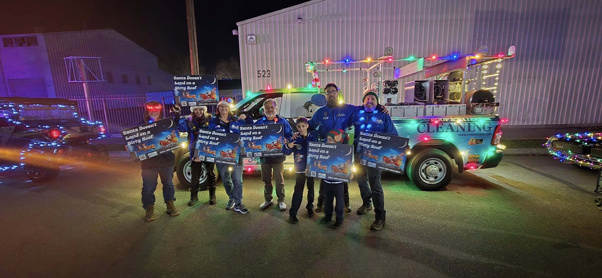 Group of people holding signs that read "Santa Doesn't Land on a Dirty Roof," wearing festive attire, in front of a brightly lit truck promoting Clearview SoftWash holiday lighting services.